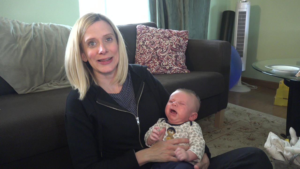 Blonde woman holding a crying baby on a living room floor.