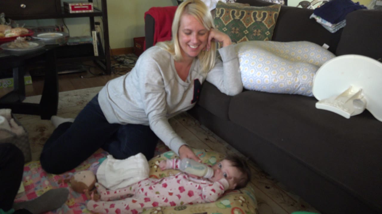 Mother kneels beside an infant and holds a bottle in a living room.