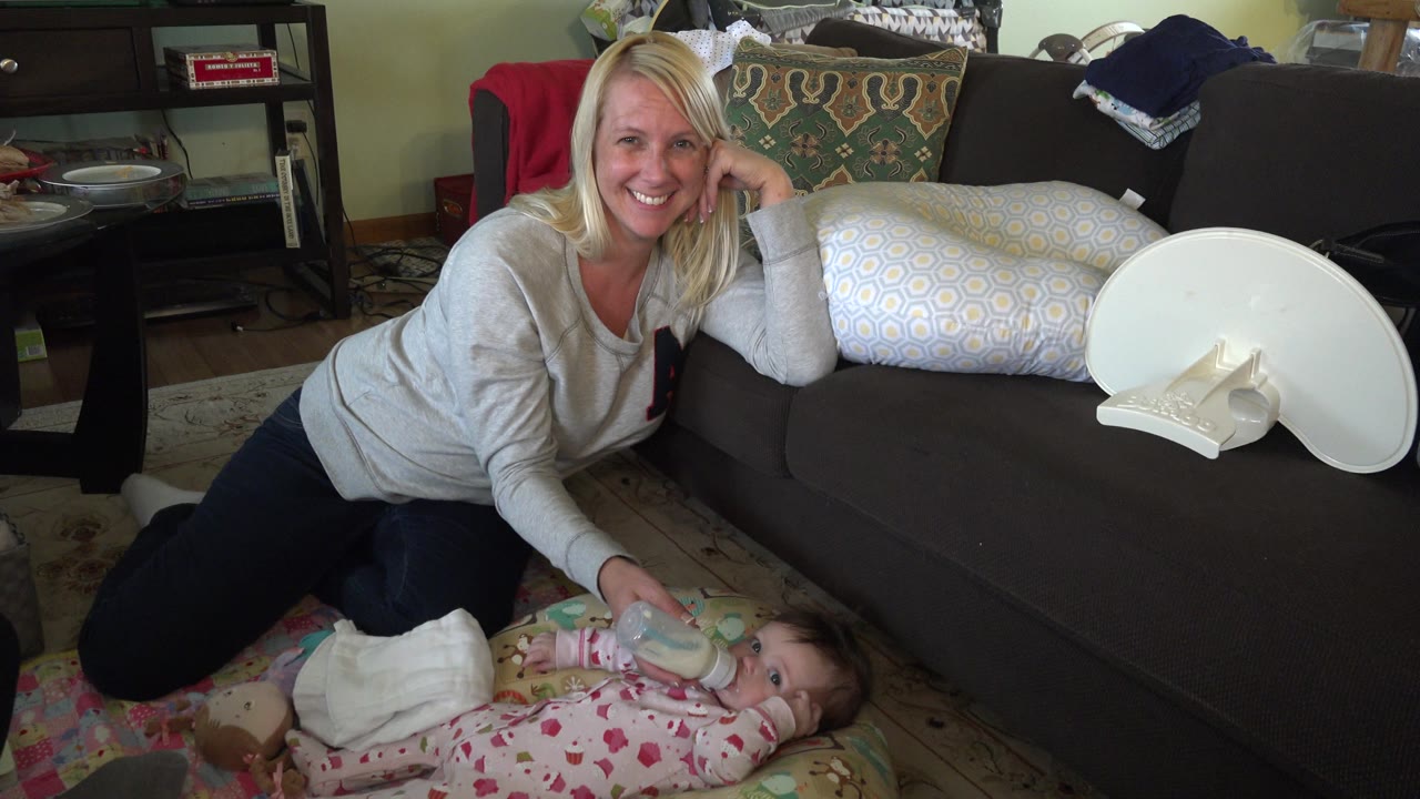 Blonde woman smiling while feeding an infant beside a couch.