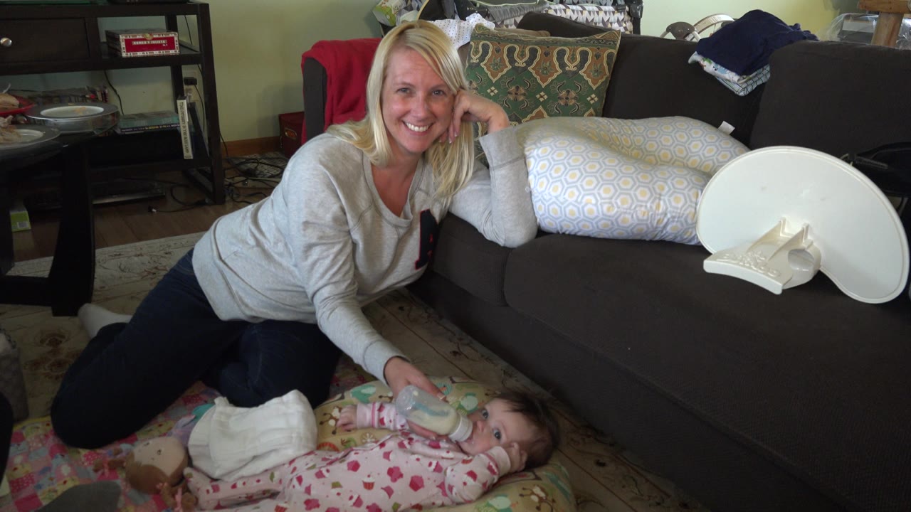 At 0:01 in Hidden Lake, Colorado, the woman remains propped beside the couch while the infant drinks from the bottle on the floor mat below her.