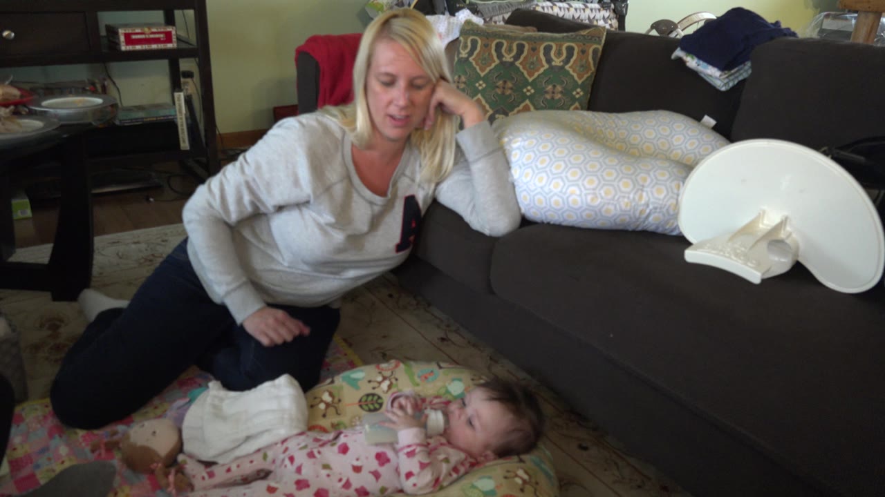 At 0:00, a blonde woman kneels near a baby lying on a patterned floor mat with a bottle in a Hidden Lake, Colorado living room.