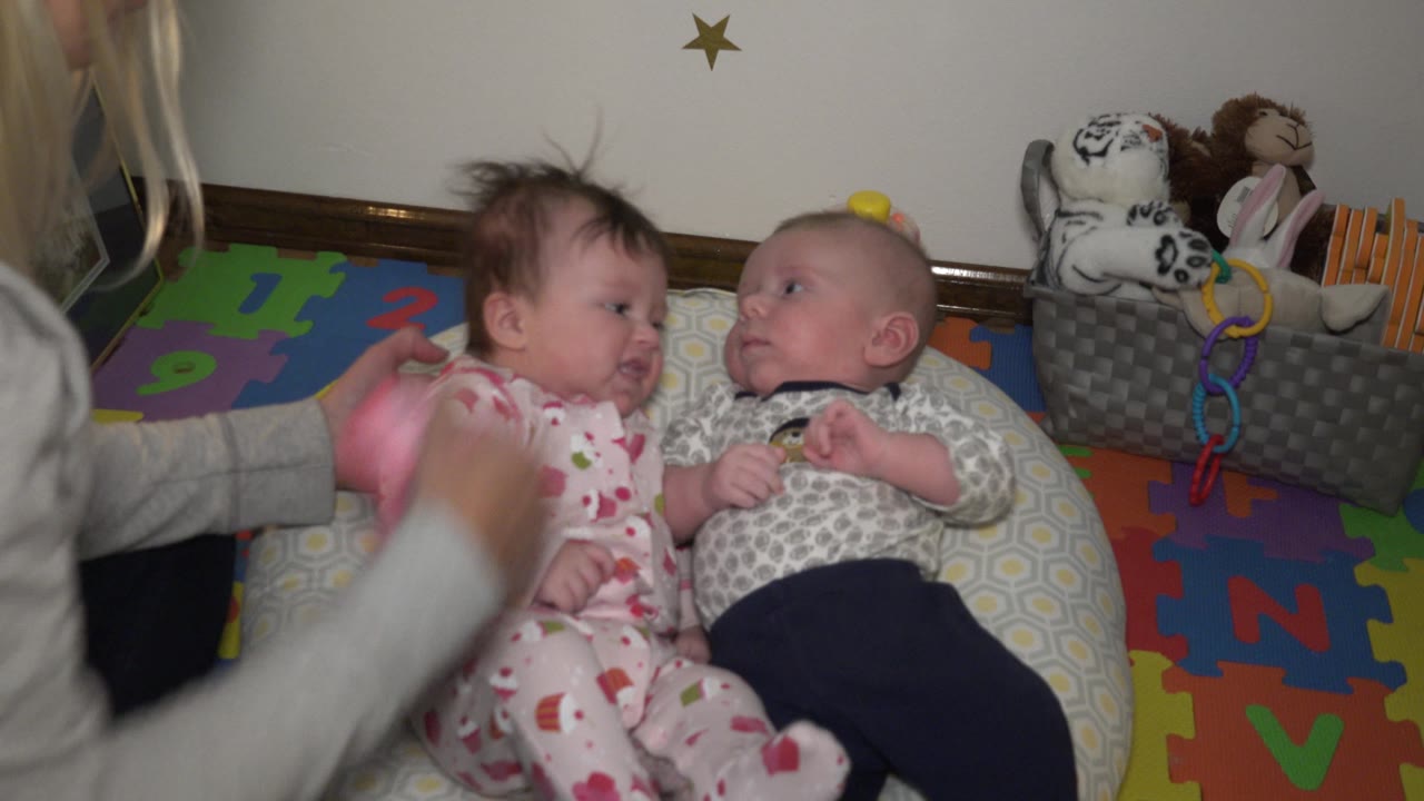 An adult arm moves out of the foreground as the baby in pink patterned pajamas turns slightly toward the baby in gray, with the alphabet mat visible around the pillow.