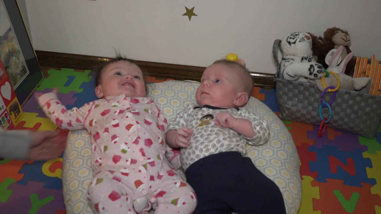 The babies remain on the floor pillows as an adult arm reaches in from the left edge, with the colorful foam mat and toy bin still visible behind them.