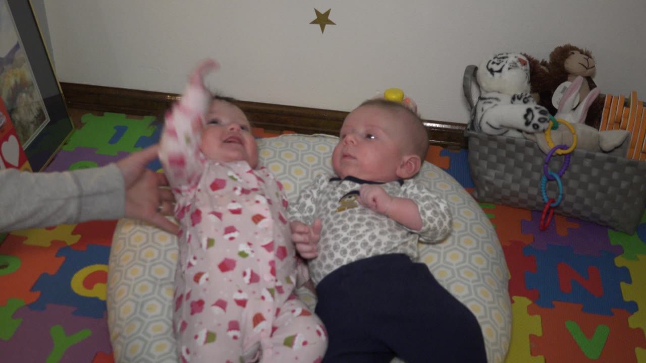 An adult hand lifts the left baby's pink-sleeved arm while the second baby looks across from a neighboring pillow in the same indoor play area.