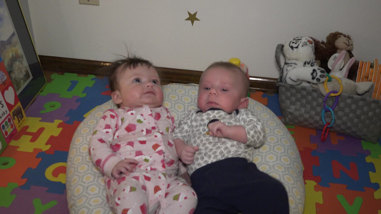 Two infants lying side by side on a patterned cushion.
