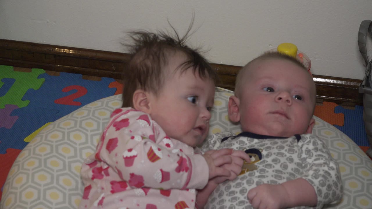 At 0:00, two babies lie side by side on a patterned cushion, with one in pink pajamas leaning toward the other while their hands touch.