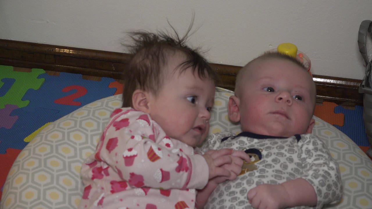 At 0:01, the baby in pink pajamas faces the baby in gray clothes as both lie on the bed with their hands together near the middle of the shot.