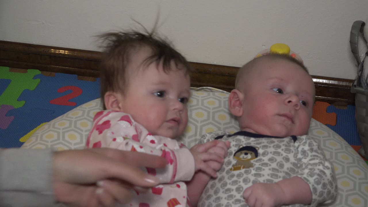 Two babies lying on a bed as an adult hand reaches into frame.