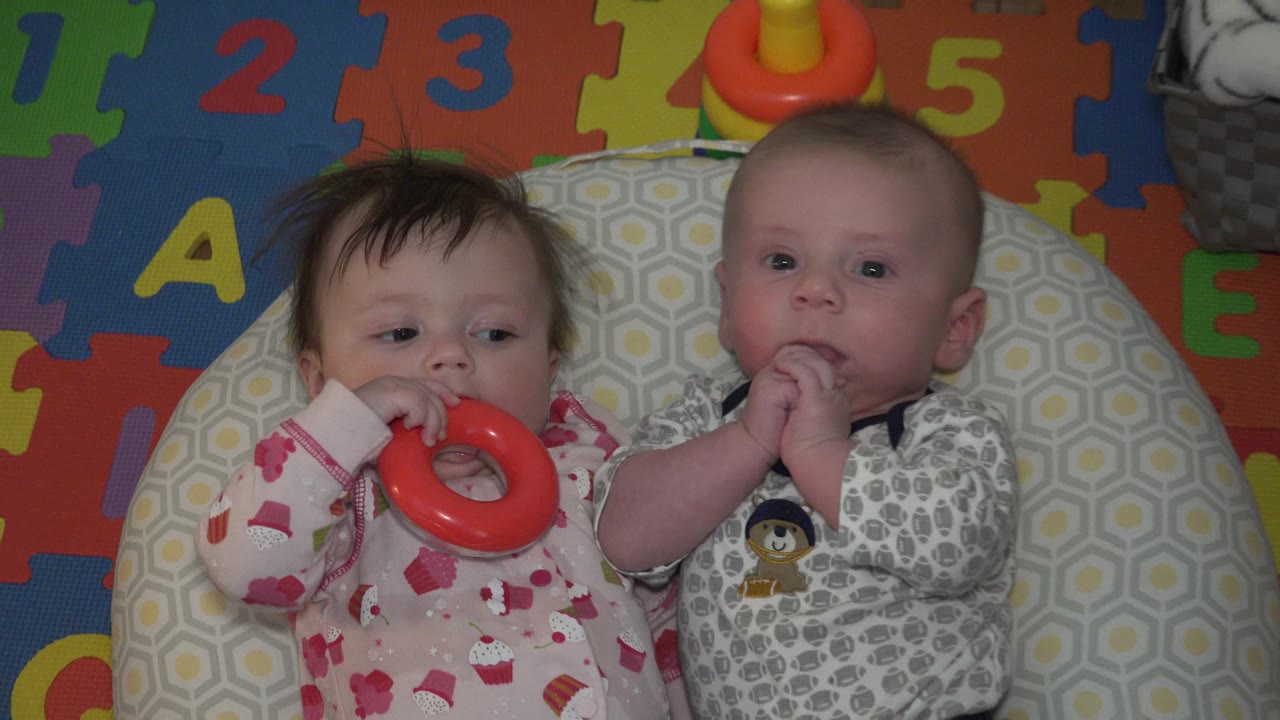 Two babies lying on a patterned pad with a red ring toy.