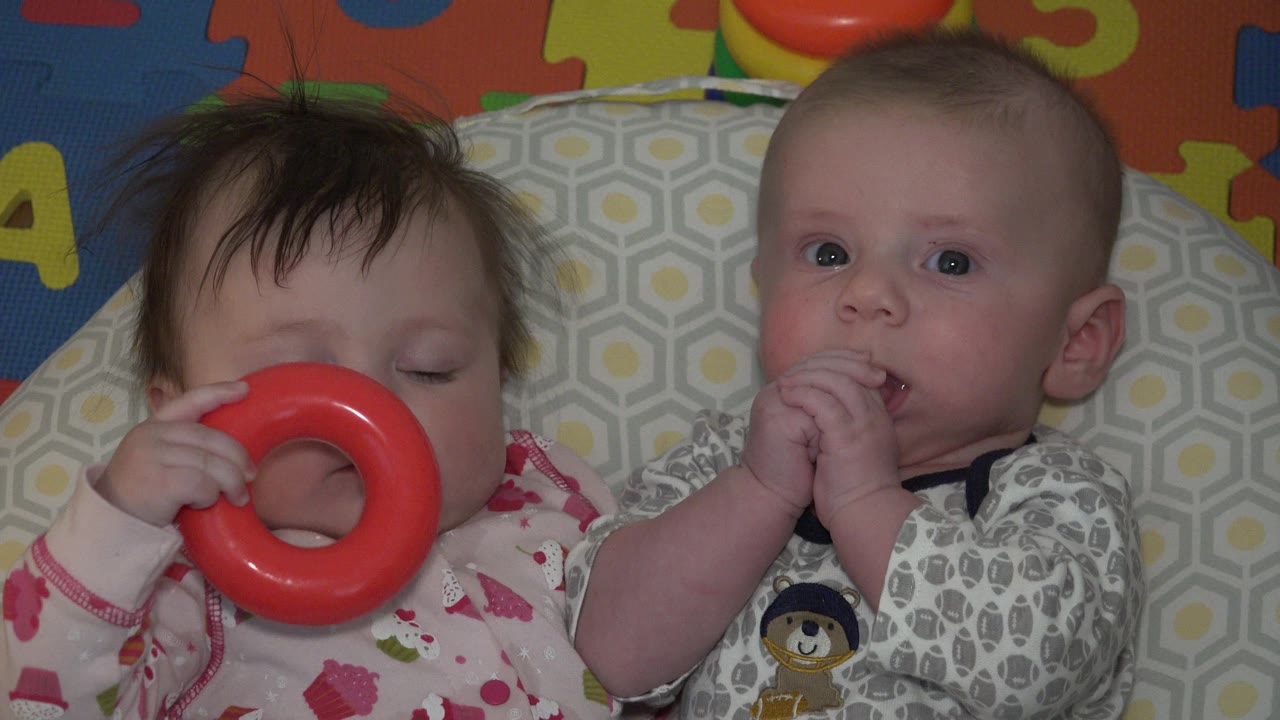Two babies lying on bedding, one holding a red ring toy.
