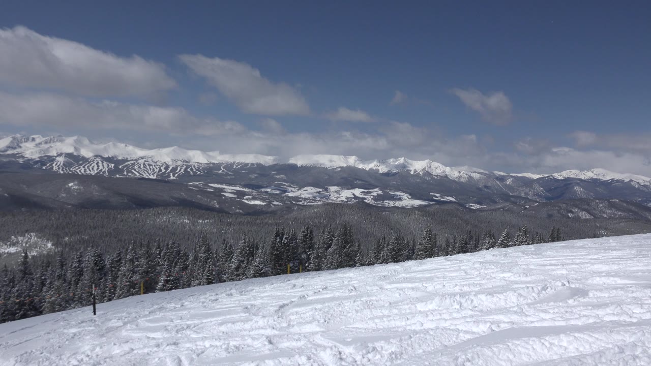 Keystone, Colorado snow field spans the foreground with forested hills and bright white mountain peaks under broken clouds, with no visible people.