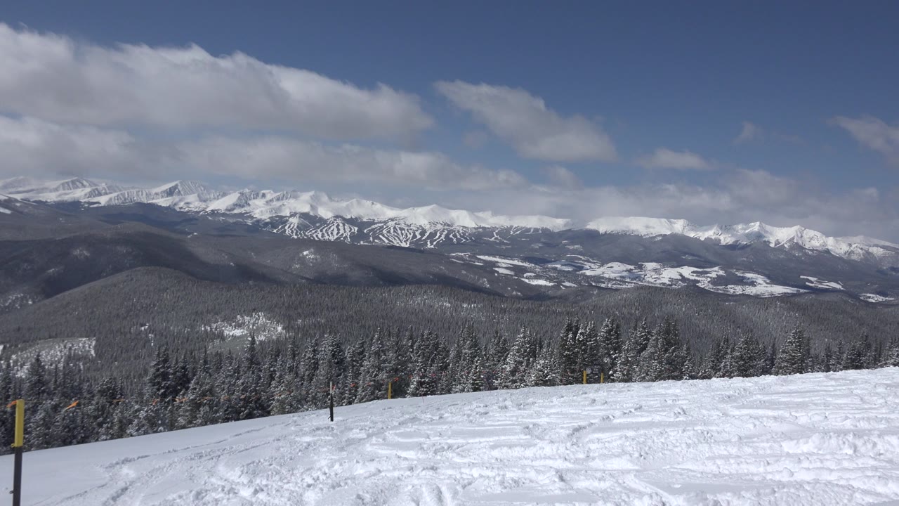 Snowy hill overlooking forested ridges and distant white mountains.