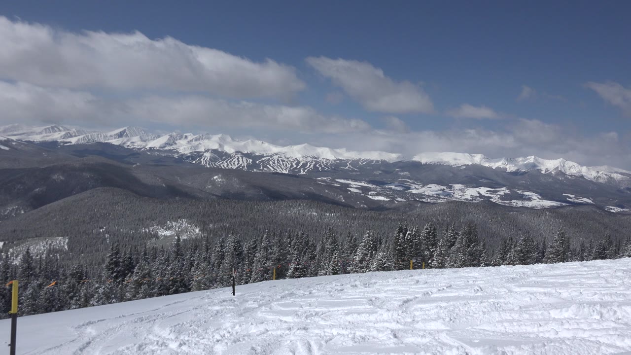 Keystone, Colorado snowfield in the foreground with a low line of frosted trees, layered dark ridges, and white mountain summits beneath a broad cloud band, with no visible people.