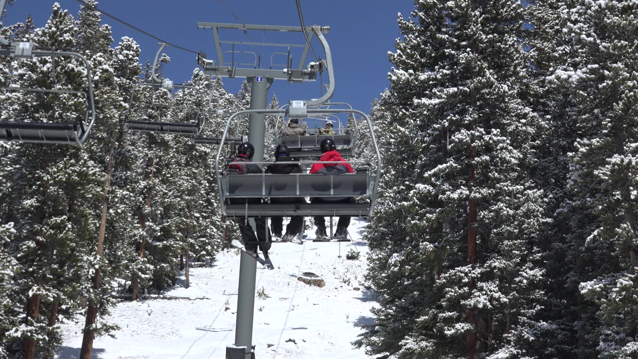 The chair carrying three skiers sits near the center of the Keystone, Colorado frame, with dense snowy pines behind it and lift cables overhead.