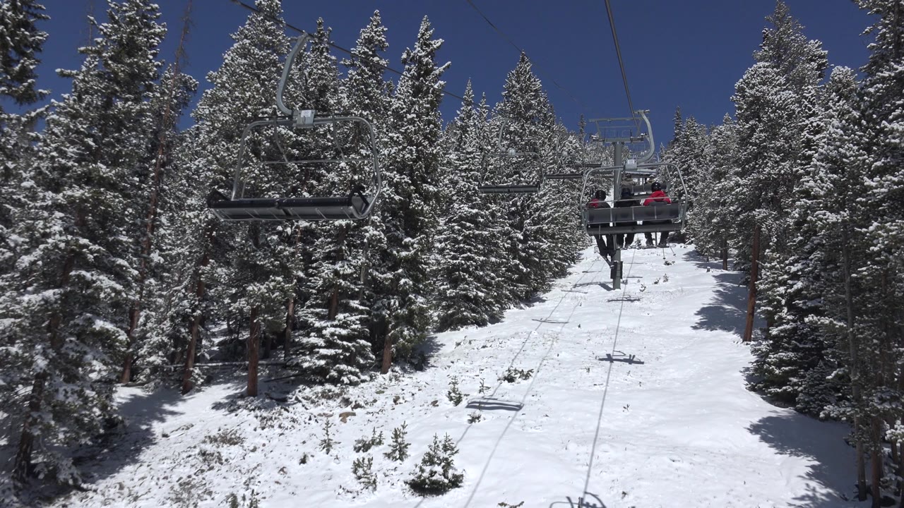 A chairlift crosses the upper part of a snow-covered Keystone slope, with evergreen trees packed on both sides and distant riders partly visible on a chair.