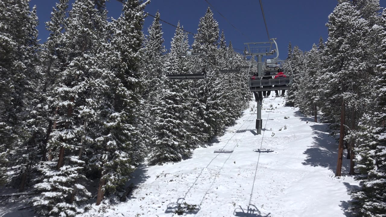 Keystone, Colorado ski lift chairs move through the upper frame over a white slope bordered by snow-dusted evergreens, with distant people on a chair.