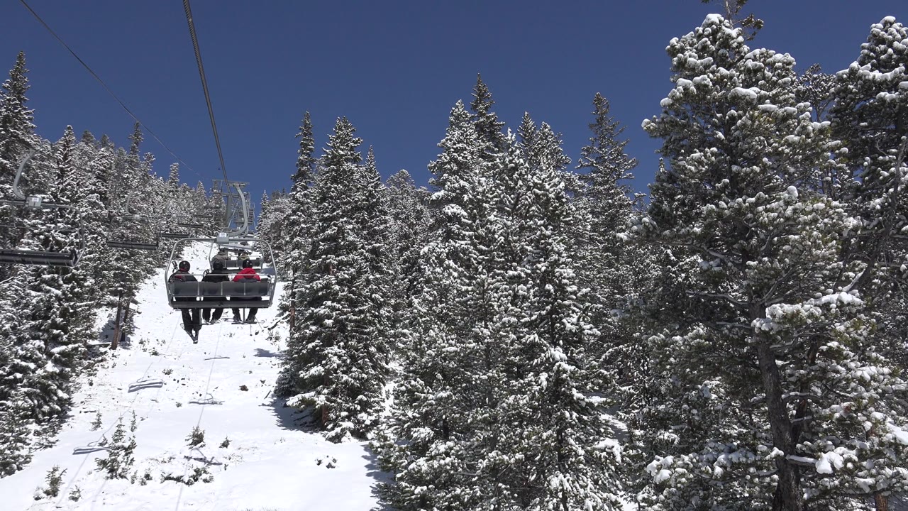 At 0:01, the chairlift is centered between snow-laden evergreens in Keystone, Colorado, with three bundled riders visible from behind under the cable line.