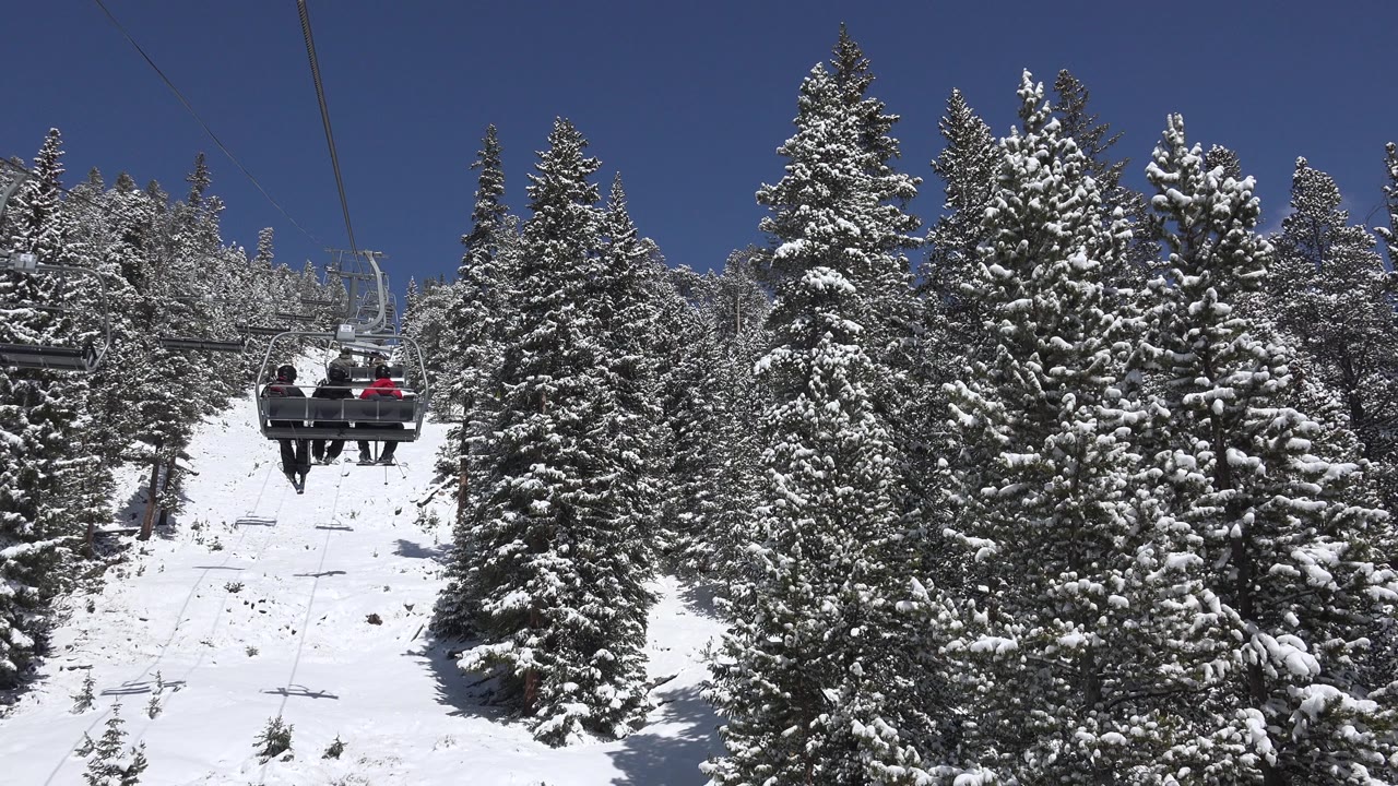 In Keystone, Colorado, bundled riders on a chairlift sit near the upper left of the snowy lift corridor, with tall evergreen trees filling the center and right side.