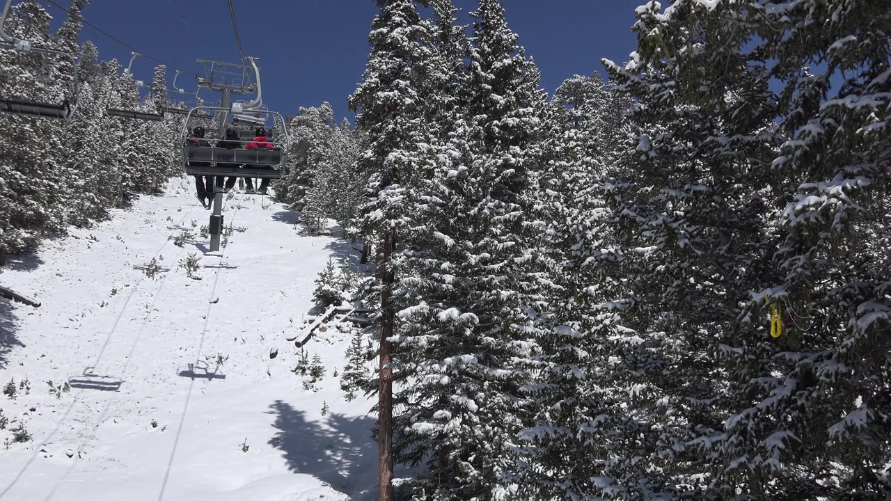 The Keystone ski lift remains centered over the snow-covered slope, with dark evergreen trees on both sides and distant chair riders visible against the blue sky.