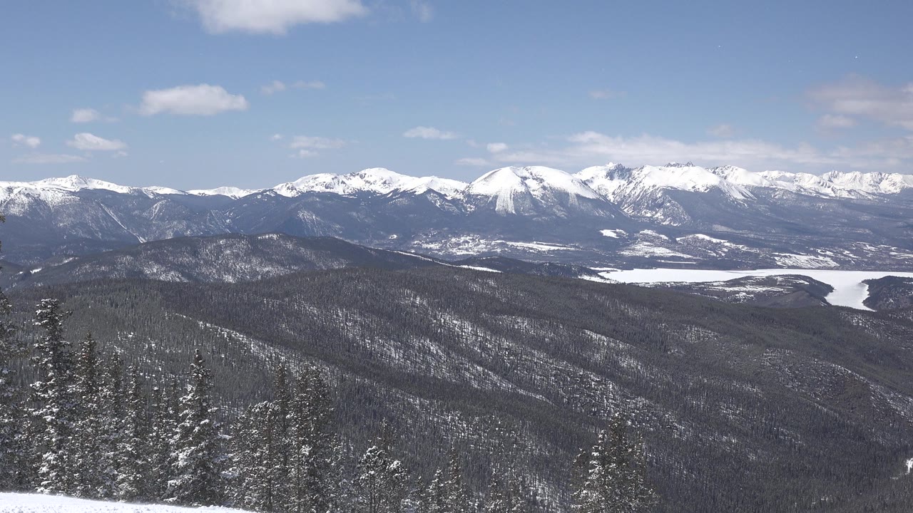 Keystone, Colorado winter vista with bright snow on distant peaks, dark rolling forest below, sparse clouds, and no visible people.