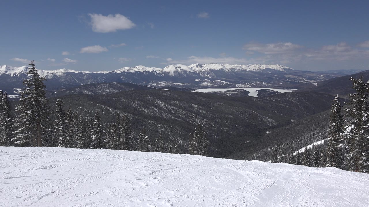 Keystone, Colorado mountain view with a bright snowfield in front, tall evergreens near the center-right, layered dark ridges, distant white peaks, and no visible people.