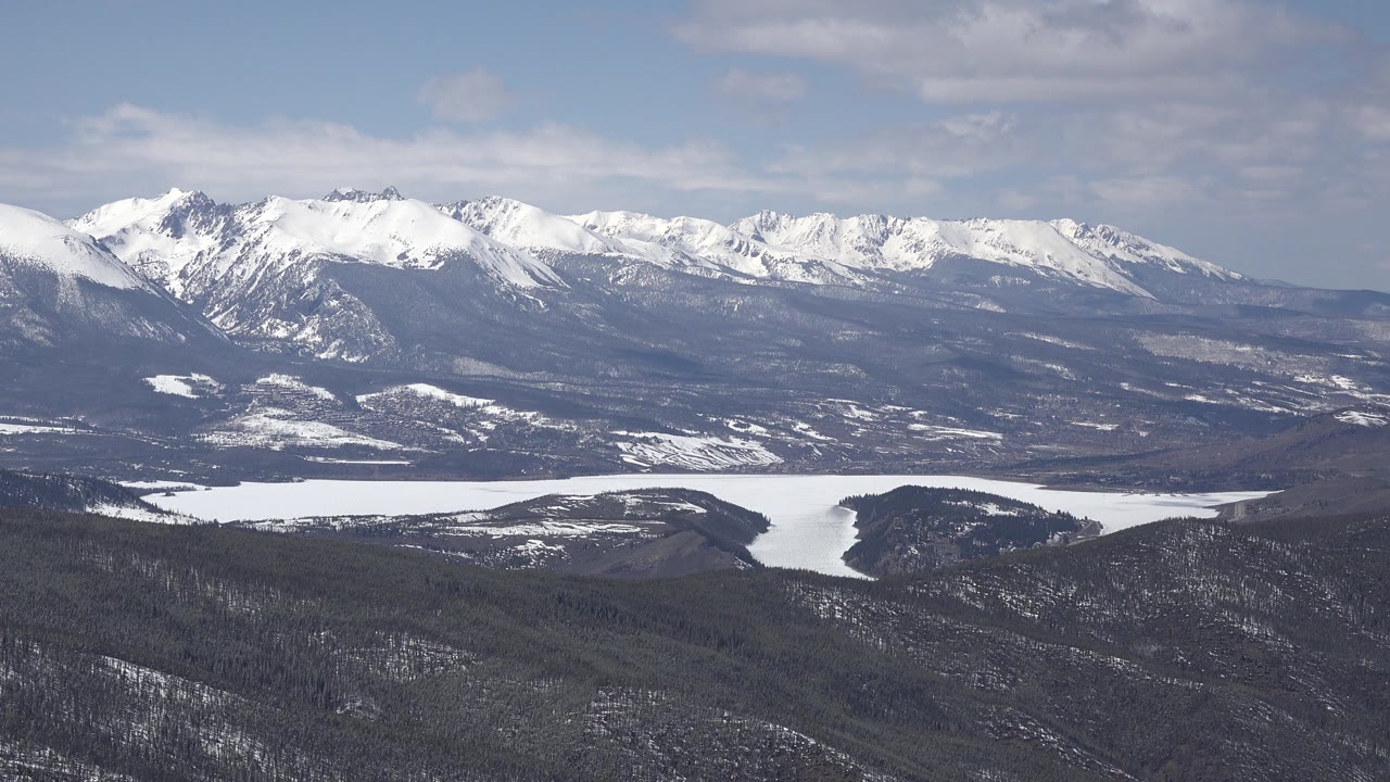 Wide view of snow-capped mountains near Keystone, Colorado, with a pale valley water shape below, dark forested hills in front, and no visible people.
