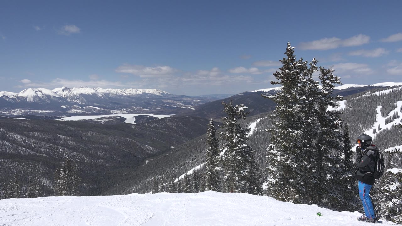 Snowy mountain overlook with evergreens and a bundled adult at right.