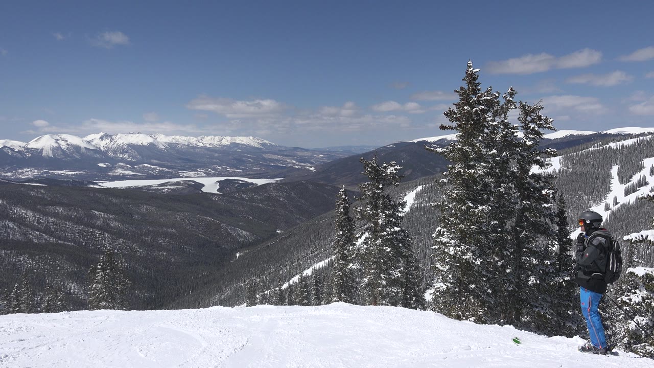 Keystone, Colorado summit-style view showing a bright snow ledge, evergreen trees, layered dark slopes, distant white peaks, and a bundled adult partly near the right edge.