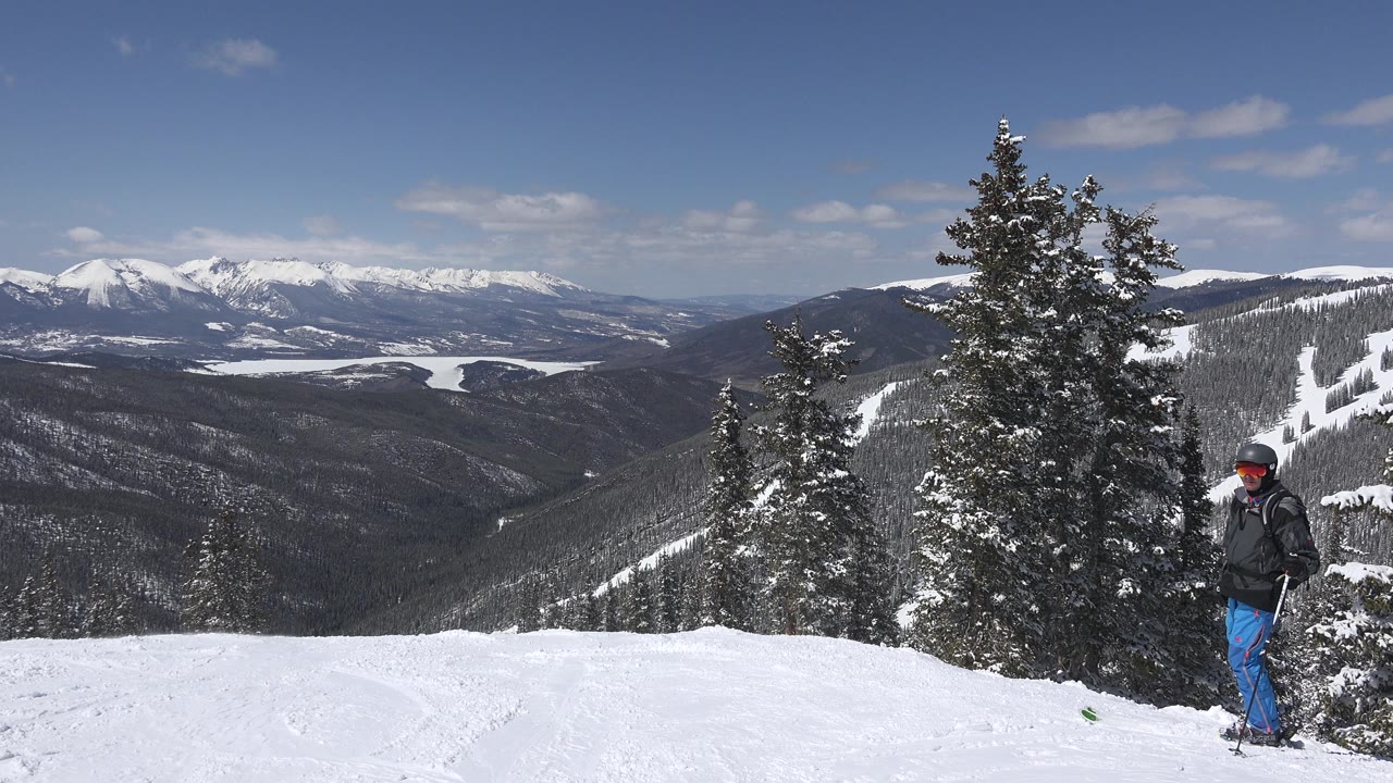 Man in blue pants standing on snow near evergreen trees and distant mountains.