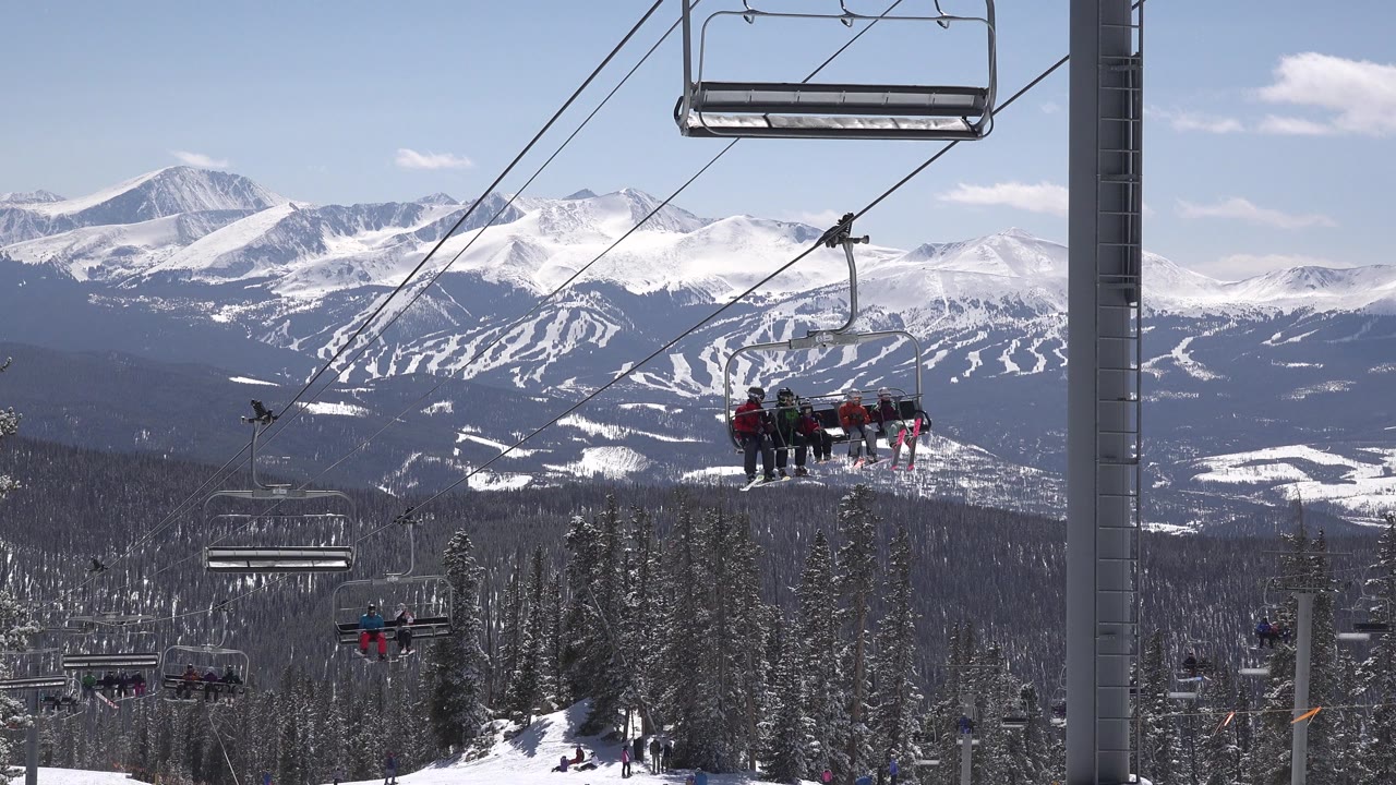 Bundled people ride a chairlift near the center of the frame in Keystone, Colorado, while empty chairs, diagonal cables, snowy trees, and mountain slopes fill the view.