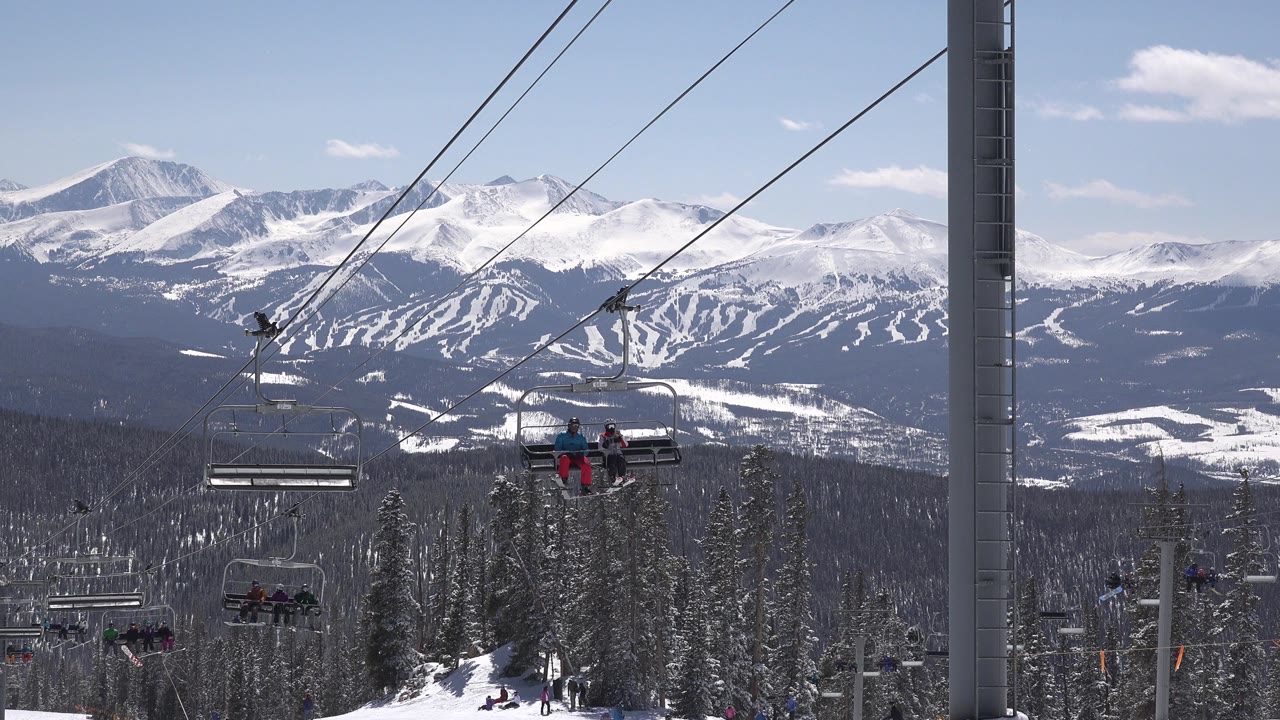 Chairlifts over snowy evergreens with mountain ski runs in the distance.