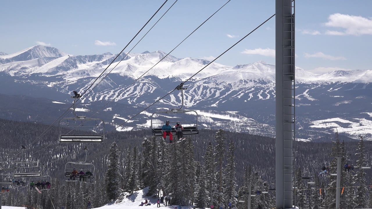Two visible riders sit on a Keystone, Colorado chairlift, with a lift tower, snowy trees, distant ski runs, and mountains behind them.