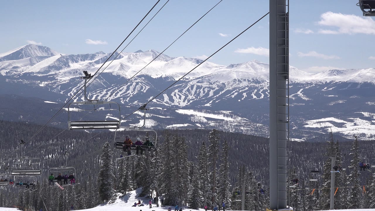 A Keystone, Colorado chairlift line crosses snowy evergreens, with several riders visible on distant open chairs and white mountain ridges in the background.