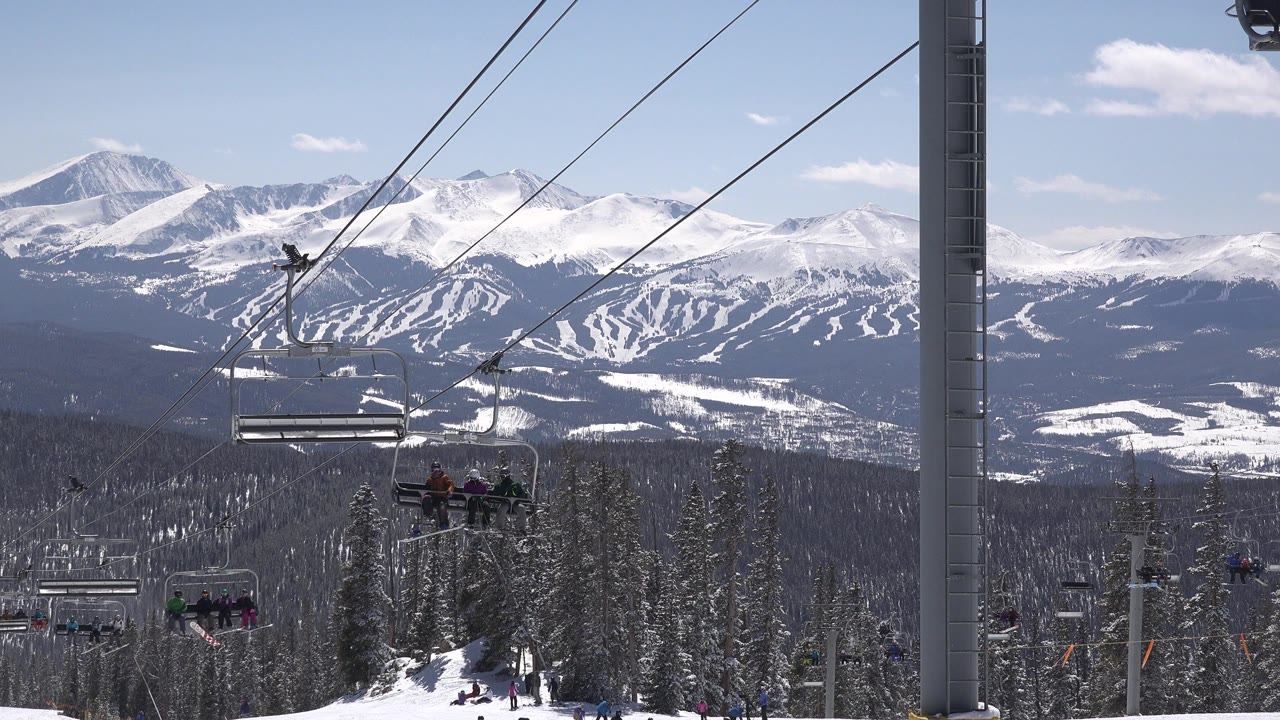 Riders on open ski lift chairs move through the Keystone, Colorado frame, with diagonal cables above a dark evergreen slope and bright snowy peaks beyond.