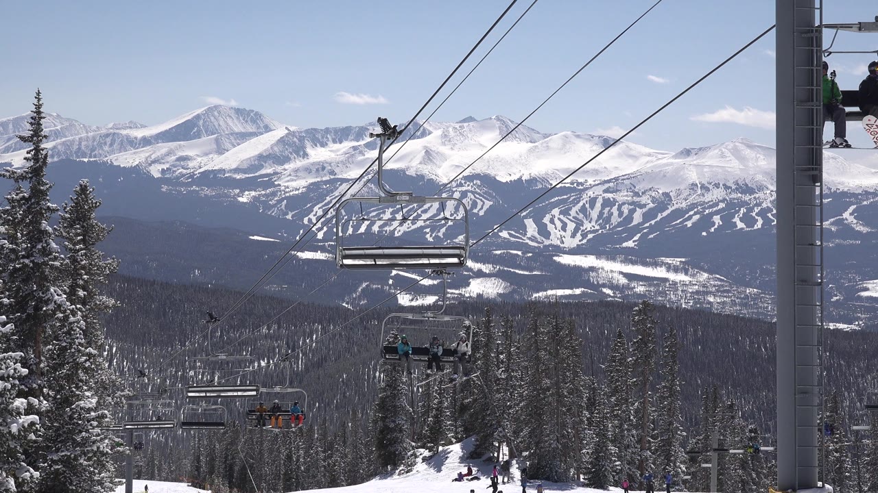 A gray lift tower stands near the center as chairlift cables cross the sky over Keystone, Colorado, with snowy trees below and distant riders on the chairs.