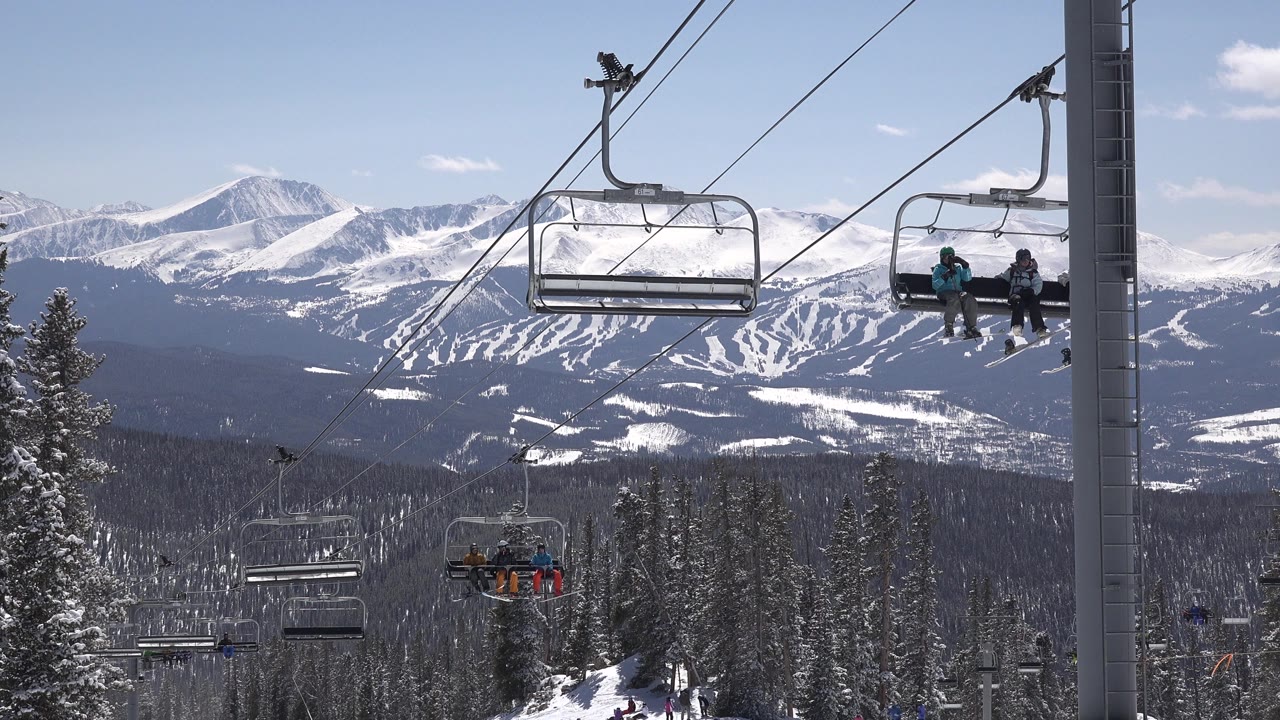 Chairlifts over snowy pine forest with mountain ski runs in the background.
