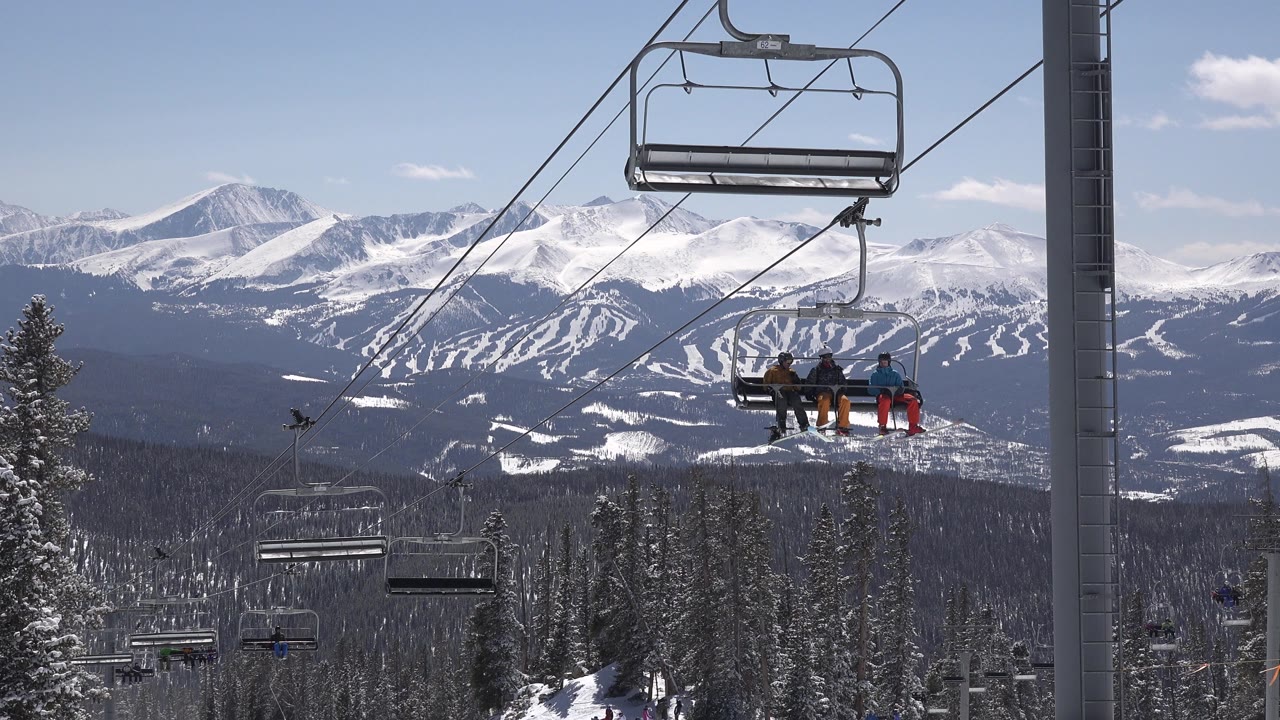 Chairlift riders above snowy forest with mountain peaks behind them.