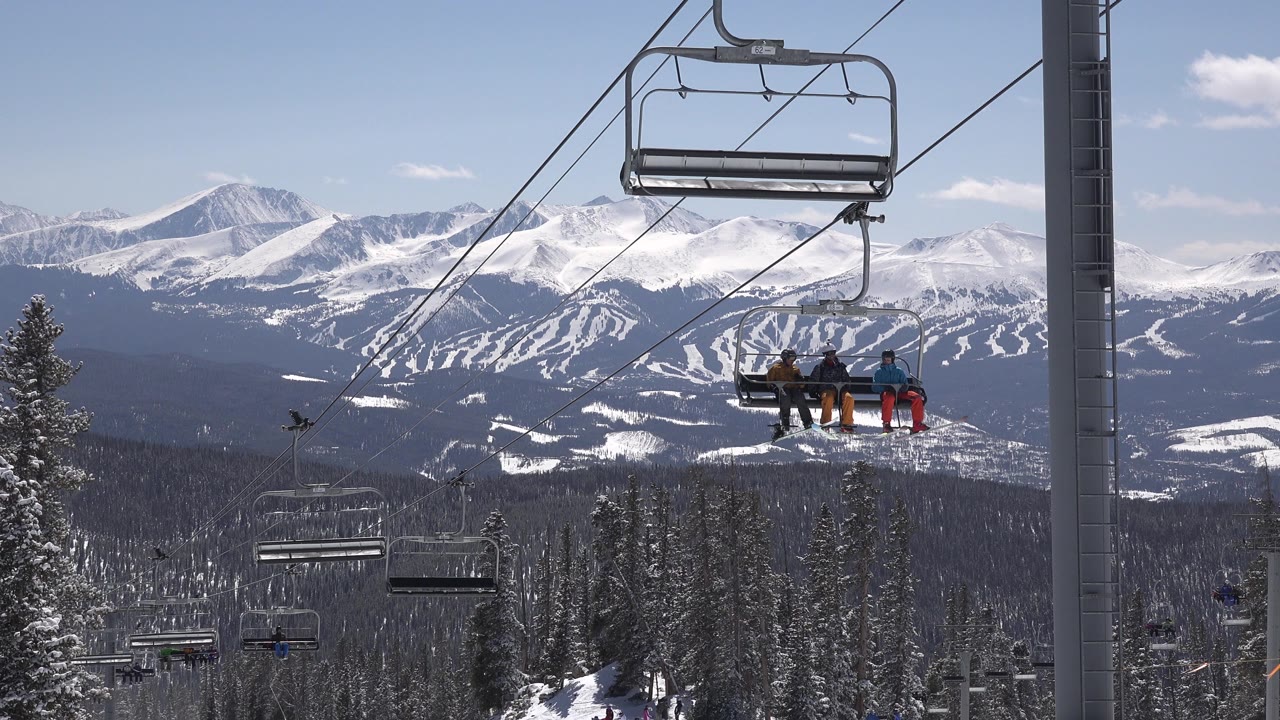 Riders on a chairlift move across the Keystone, Colorado mountain view, framed by diagonal lift cables, a gray lift tower, dark forest, and white ridges beyond.