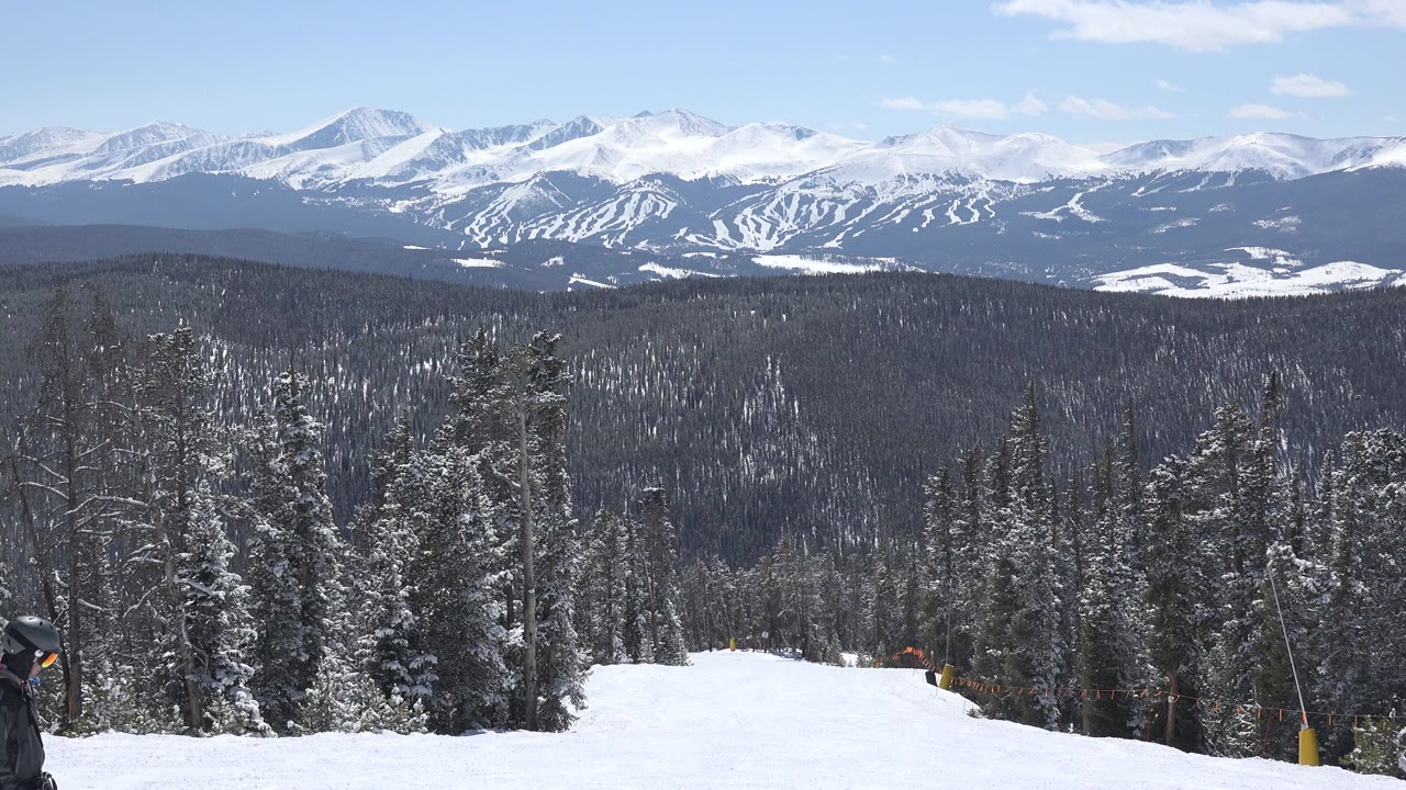 Keystone, Colorado winter view from a snowy slope toward evergreen woods and a distant mountain range, with a partial skier at the far left edge.