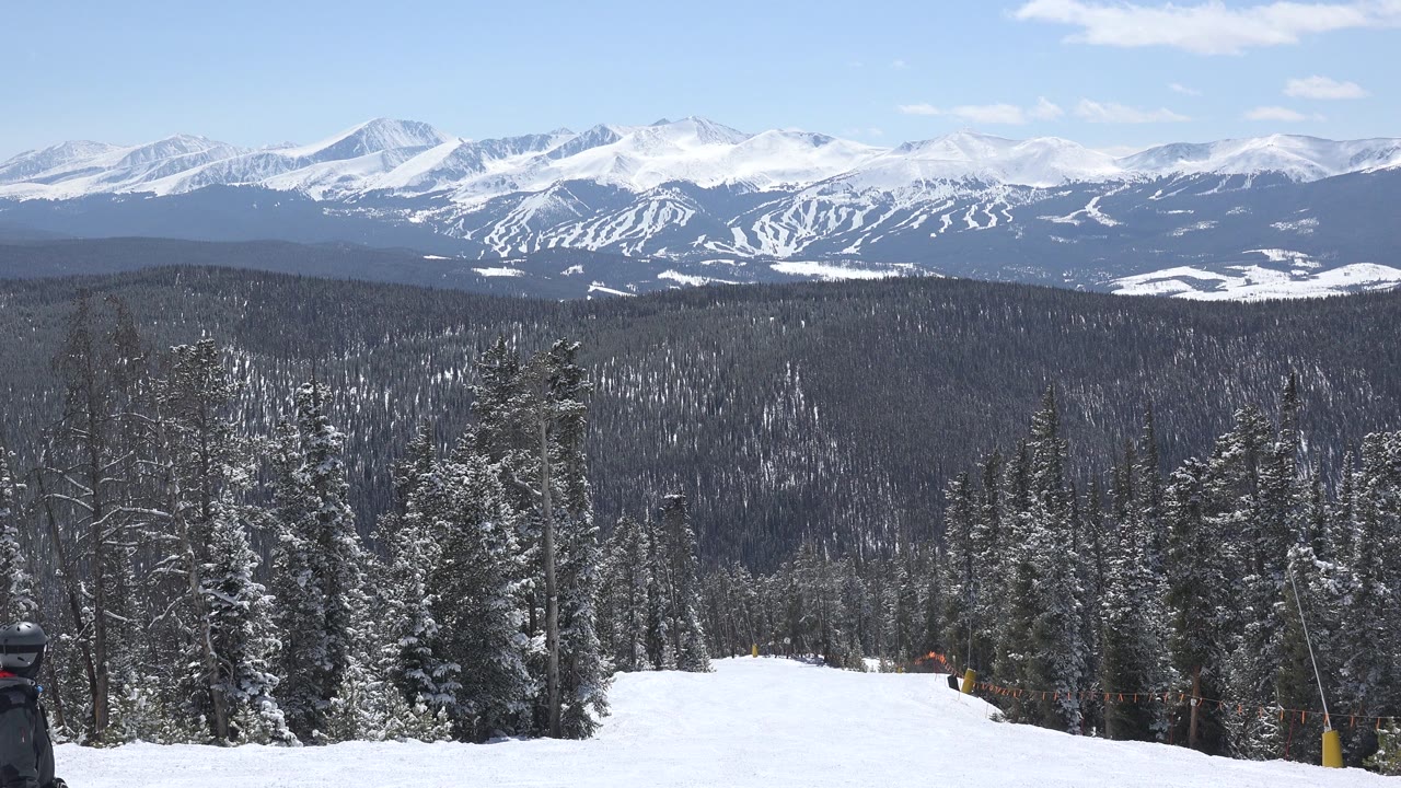 Snowy mountain slope bordered by evergreen forest and distant peaks.