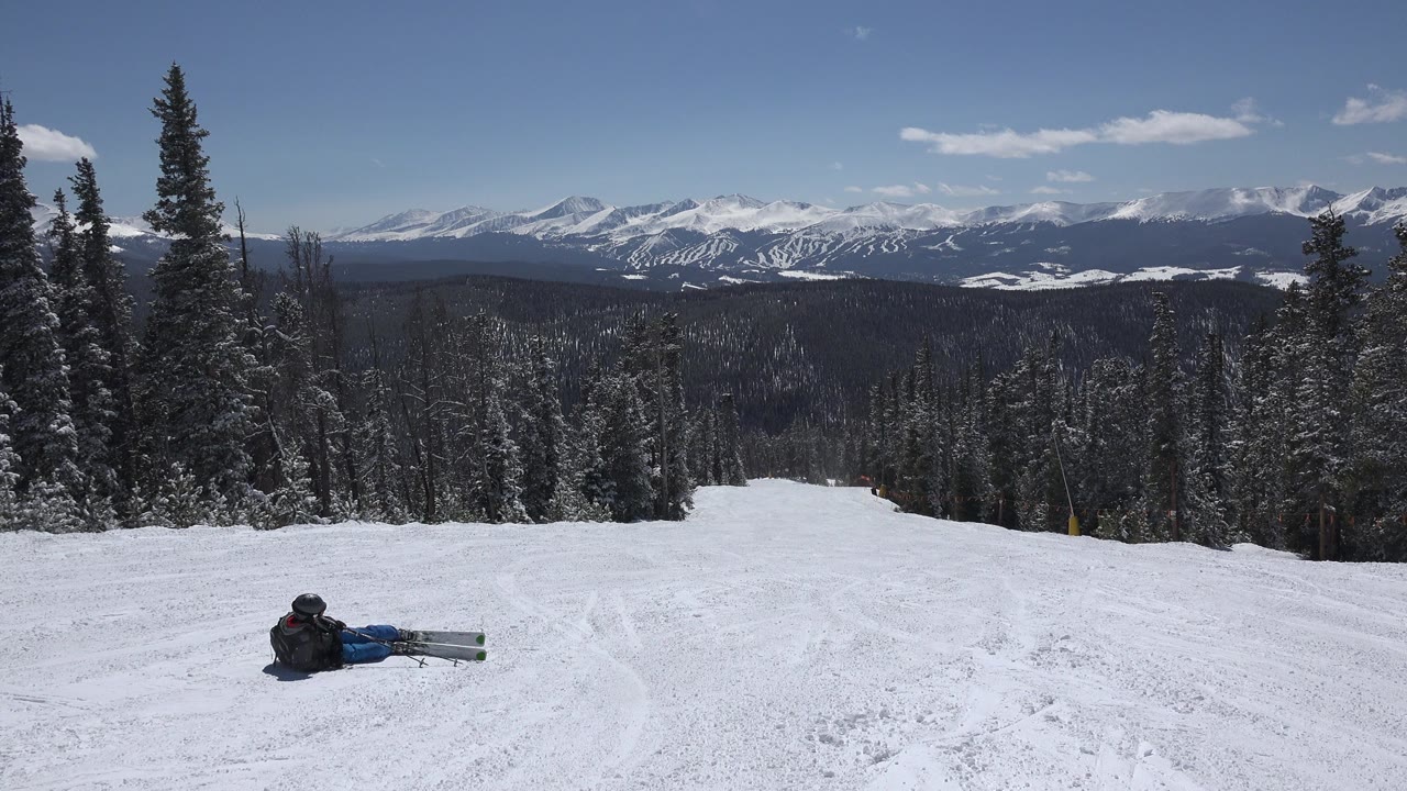 Snowy ski slope with seated skier, evergreen trees, and distant mountains.