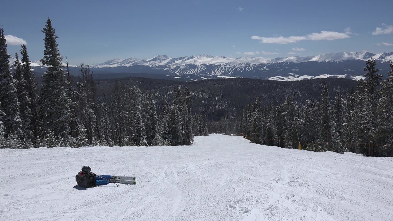 A Keystone, Colorado winter slope remains framed wide, with the seated skier near the lower center-left, tracks in the snow, dark forest edges, and distant white mountain ridges.