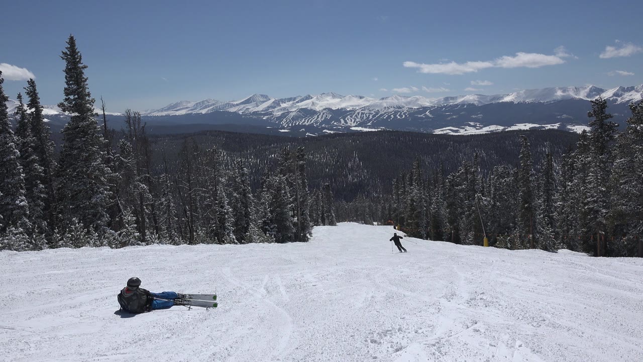 Snowy ski slope with pine trees, distant mountains, and two people on the run.