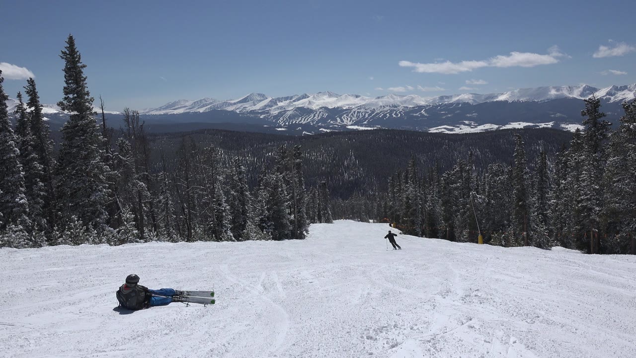 Keystone, Colorado ski run framed by pine trees, with the seated person still low in frame and the distant skier moving farther along the open slope.