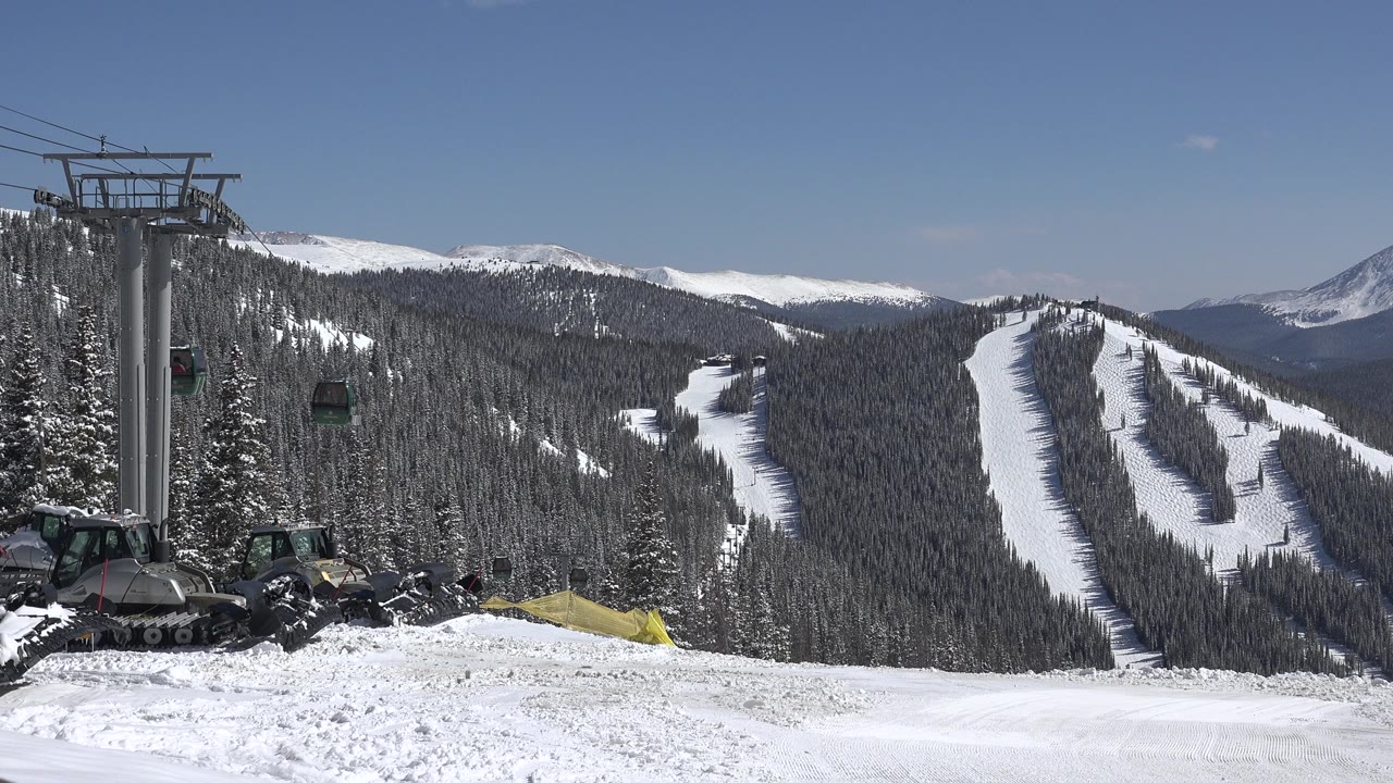Opening frame of Keystone, Colorado ski terrain with a lift tower and gondola cars on the left, snow vehicles in the foreground, wooded slopes, distant ski runs, blue sky, and no visible people.