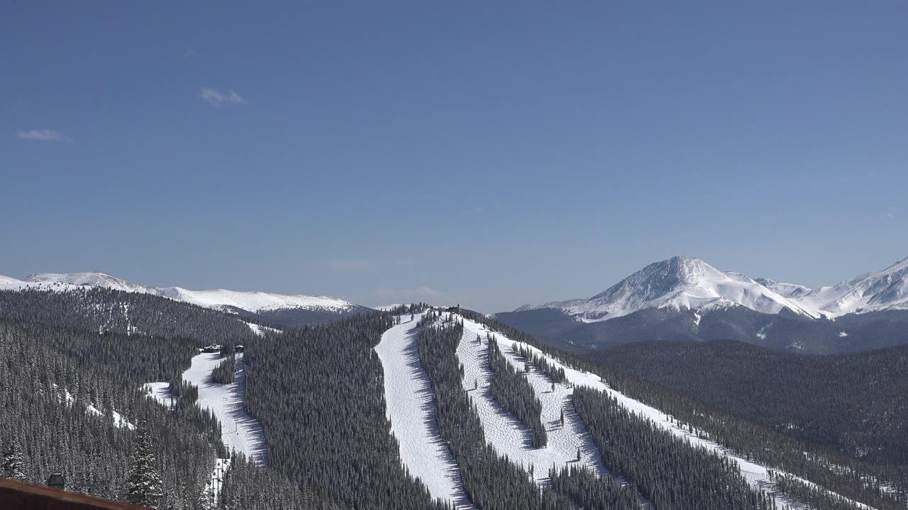 Snowy mountain ridge with ski runs, evergreen forest, distant peaks, and blue sky.