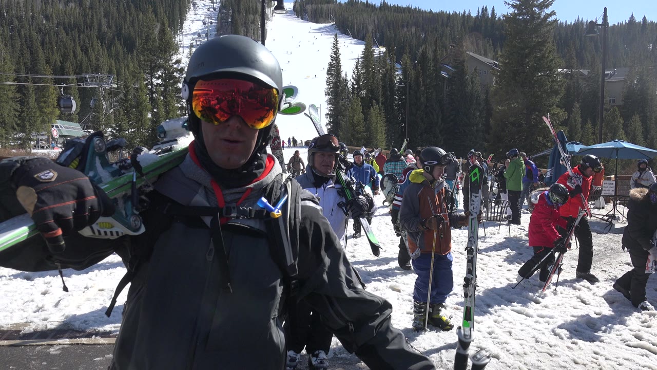 At Keystone, Colorado, the foreground skier remains close to the lens with one gloved hand raised, while people carrying skis move across the sunlit snow behind him.