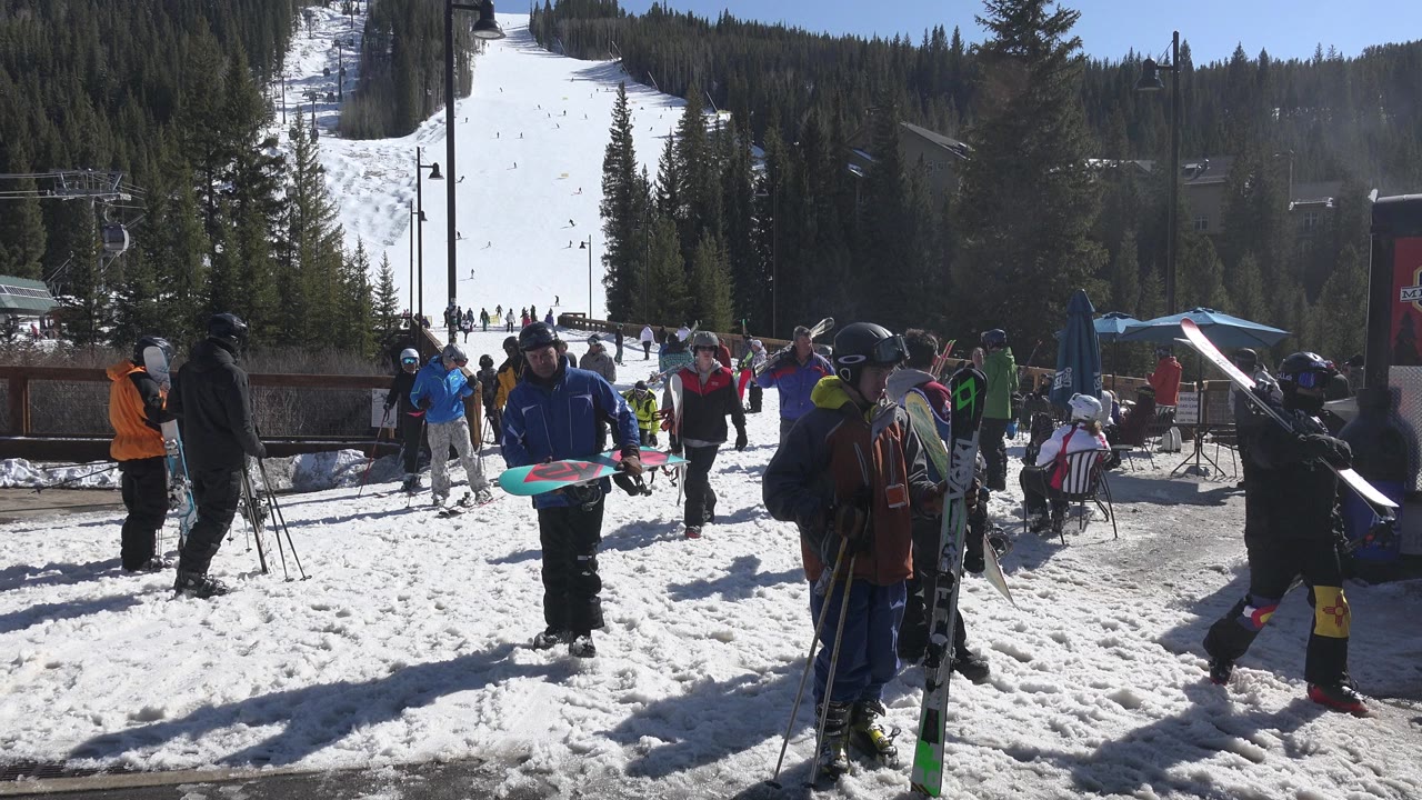 A snowboarder carrying a board passes near skiers in Keystone, Colorado, while other riders wait on the snow below the tree-lined slope.