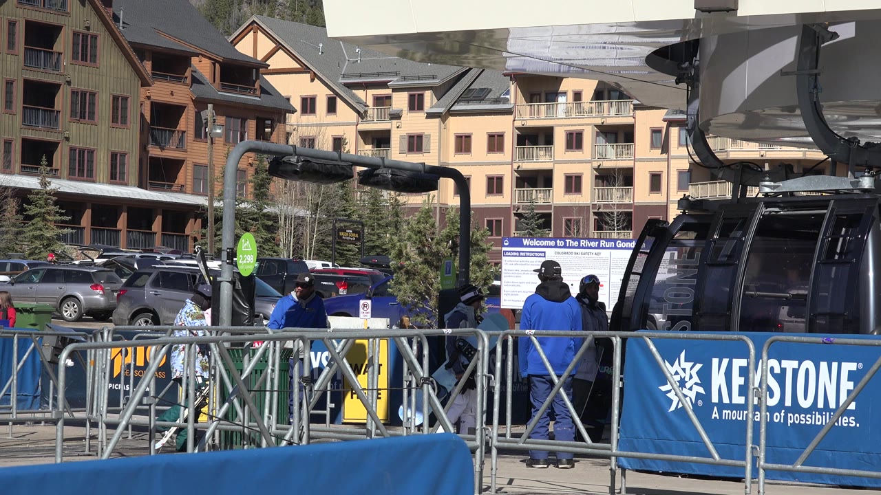 The Keystone, Colorado gondola cabin remains near the right side while visitors in dark and blue jackets stand behind the metal railings in bright sun.