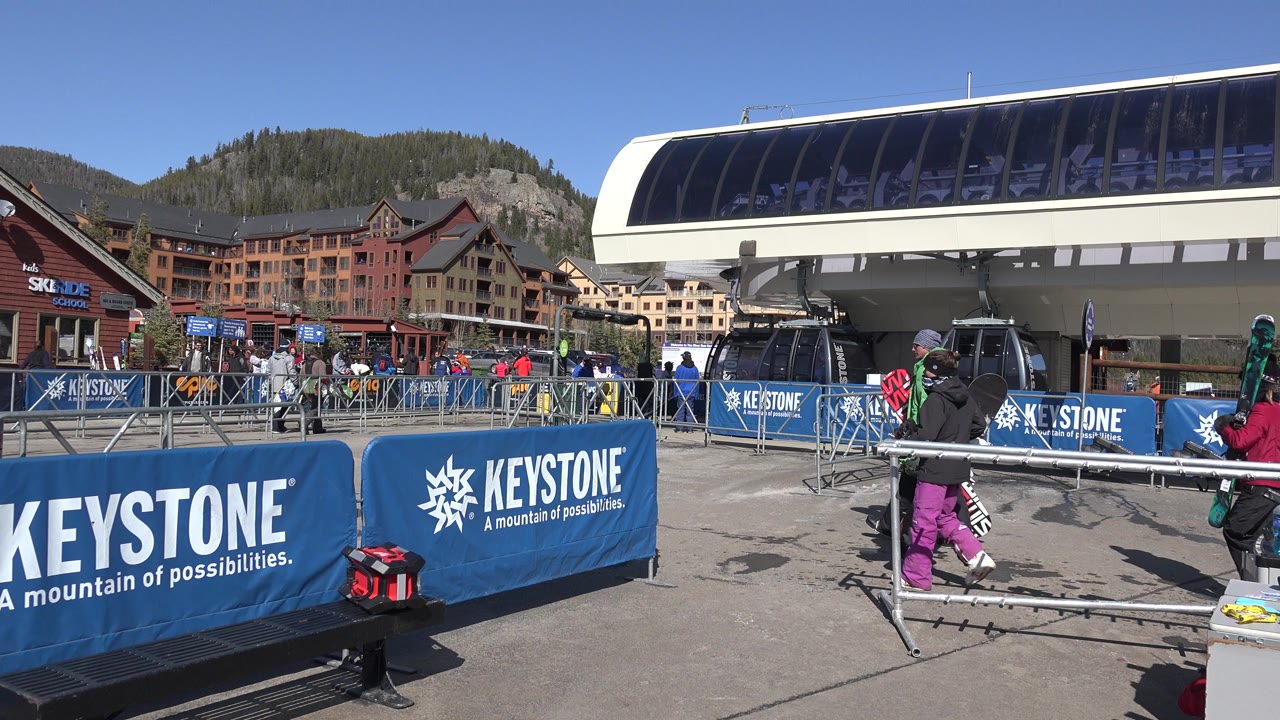 Two snowboarders carrying boards move along the Keystone chairlift queue, with blue branded barriers and the lift terminal filling the foreground and background.